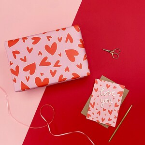 Pu&ograve; includere: Una composizione piatta con oggetti a tema San Valentino. Un regalo avvolto con sfondo rosa e motivo a cuore rosso &egrave; accanto a un biglietto d'auguri con la scritta "love you". Sono presenti anche una penna e delle forbici dorate.