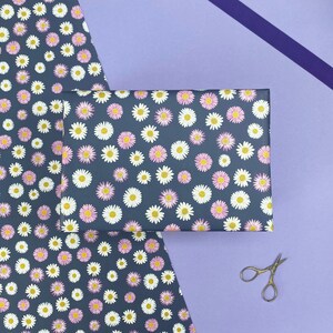 Pode incluir: Uma folha de papel de embrulho azul escuro com um padr&atilde;o de margaritas brancas e rosa. O papel est&aacute; dobrado ao meio e um par de tesouras prateadas est&aacute; sobre uma superf&iacute;cie roxa.