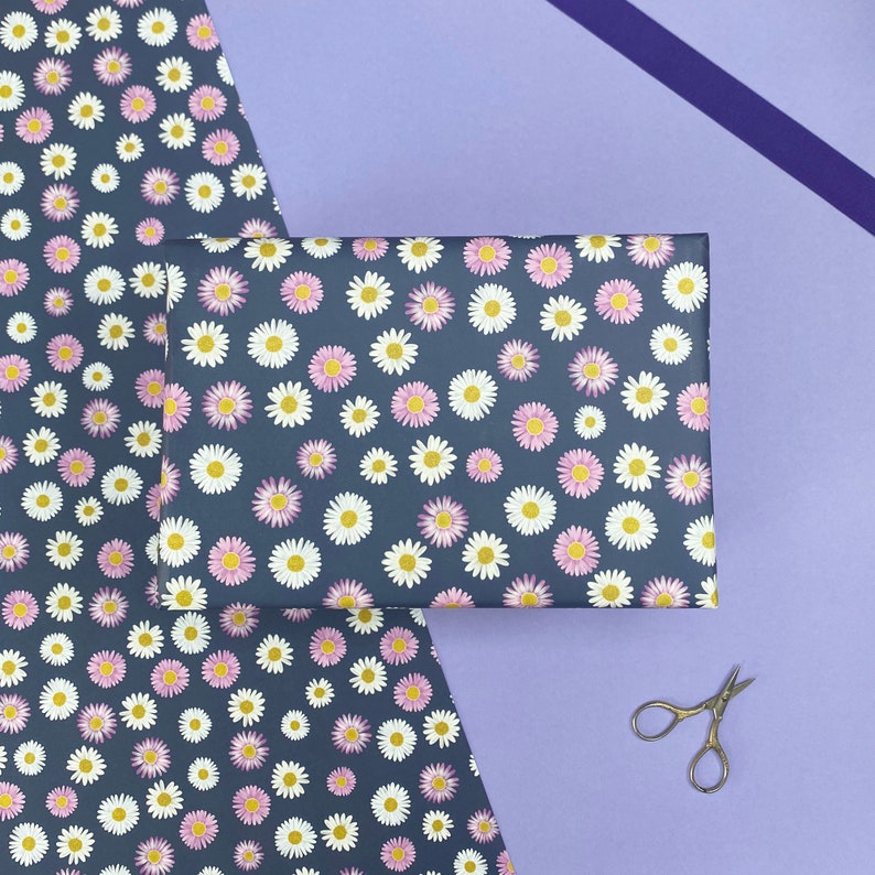 Pode incluir: Uma folha de papel de embrulho azul escuro com um padr&atilde;o de margaritas brancas e rosa. O papel est&aacute; dobrado ao meio e um par de tesouras prateadas est&aacute; sobre uma superf&iacute;cie roxa.