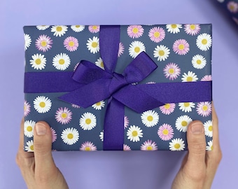 May include: A gift box wrapped in gray wrapping paper with a white and pink daisy pattern. The box is tied with a purple ribbon and bow.