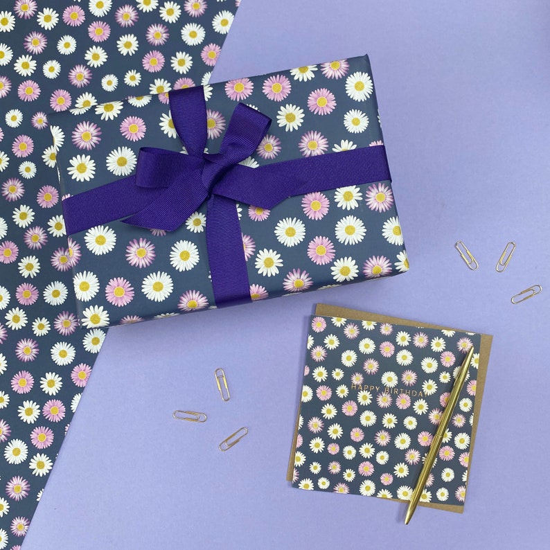 Pode incluir: Um presente embrulhado em papel de embrulho azul escuro com um padr&atilde;o de margaridas brancas e rosa. O presente &eacute; amarrado com uma fita roxa. Um cart&atilde;o com o mesmo padr&atilde;o de margarida e o texto "Feliz anivers&aacute;rio" est&aacute; sobre uma superf&iacute;cie roxa com clipes de papel dourados.