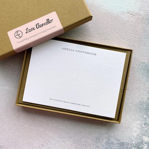 Può includere: Un set di cancelleria con una scatola marrone, un'etichetta rosa e un blocco note. L'etichetta recita "Lizzie Chancellor" e "Thoughtfully designed, lovingly illustrated." Il blocco note è stampato con il nome "AMELIA WESTBROOK" e un indirizzo.
