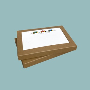 May include: Two brown cardboard boxes with a white card inside. The card has a line of four cartoon tractors in green, red, and blue colours. The text on the card reads "Grayson Chancellor".