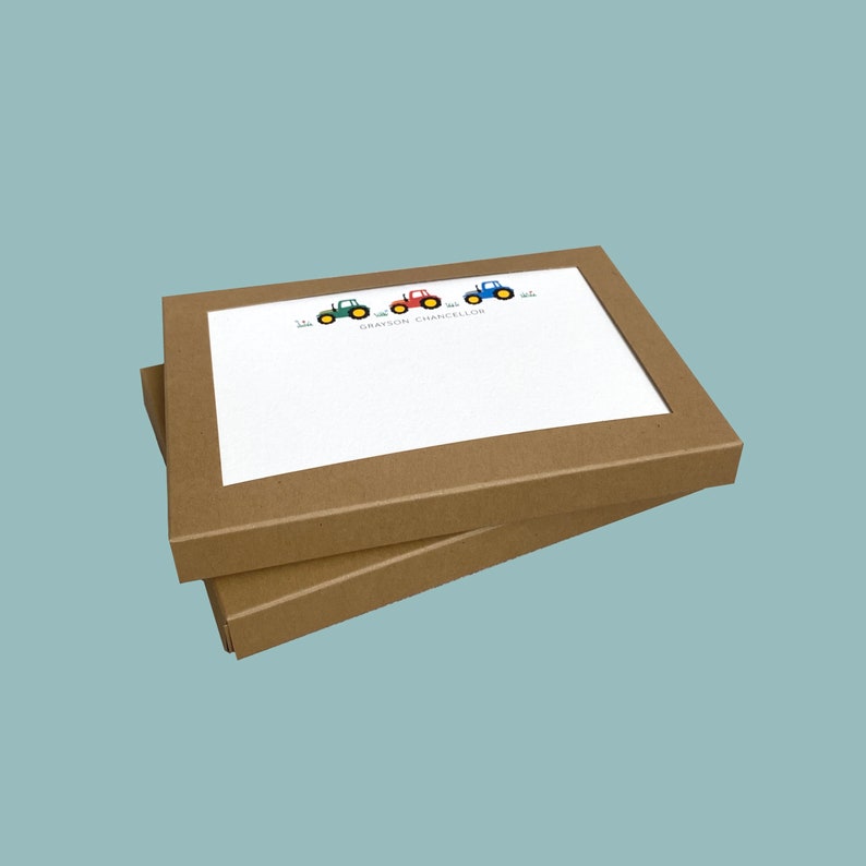 May include: Two brown cardboard boxes with a white card inside. The card has a line of four cartoon tractors in green, red, and blue colours. The text on the card reads "Grayson Chancellor".