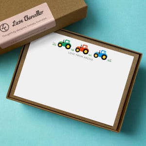 May include: A set of white stationery cards in a brown cardboard box. The cards feature a green, red, and blue tractor illustration with the text LOVE FROM ARCHIE. The box lid has a pink label with the text Lizzie Chancellor.