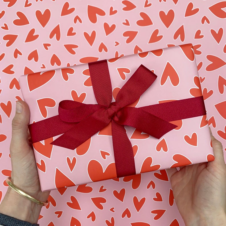 Pu&ograve; includere: Un regalo avvolto in carta rosa con un motivo di cuori rossi, legato con un nastro e un fiocco rosso scuro. Il fiocco &egrave; centrato sul regalo, creando un pacchetto accattivante.