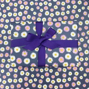 Pode incluir: Uma fita roxa amarrada em um la&ccedil;o no topo de uma caixa de presente embrulhada em papel azul escuro com um padr&atilde;o de margaridas brancas e rosa.
