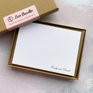 Könnte beinhalten: Ein Satz weißer Briefpapierkarten in einer braunen Schachtel. Die Karten sind leer mit den Namen "Emily and David" in schwarzer Schrift gedruckt. Die Schachtel hat ein rosa Etikett mit dem Text "Lizzie Chancellor" und "Thoughtfully designed, lovingly illustrated."