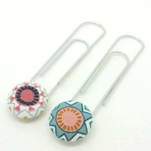 Op de afbeelding: Twee zilveren paperclips met knoppen bedekt met stof. Eén knop is wit met geometrische patronen in roze, zwart en goud. De andere knop is wit met geometrische patronen in turquoise, roze en goud.