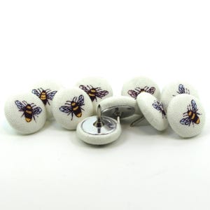 May include: A selection of decorative push pins featuring a bee motif. The pins have a white fabric covering, each adorned with a printed bee in yellow, black, and blue. The metal pin is silver. These pins are ideal for a noticeboard.