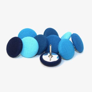 May include: A collection of blue fabric-covered push pins in various shades, including navy, teal, and light blue. The pins have a round, button-like appearance, with a silver pin extending from the center. The push pins are scattered on a white surface.