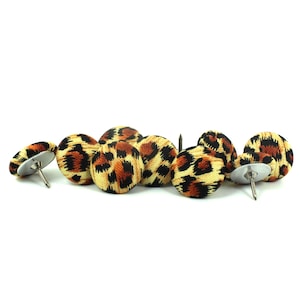 May include: A selection of leopard print upholstery tacks. The tacks have a tan fabric base with brown and black spots. The metal pins are silver.