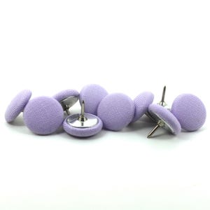 May include: A collection of round push pins featuring a light lavender fabric covering on the top surface. Each pin has a silver-toned metal base and a sharp, pointed metal needle extending from the back, designed for securing items to surfaces.