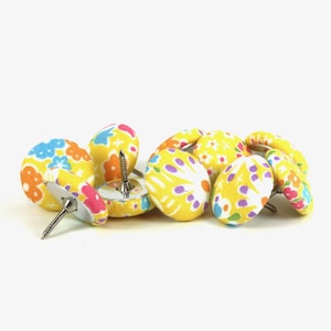 May include: A selection of decorative upholstery tacks with fabric-covered heads. The tacks have a yellow base with colourful floral patterns in pink, blue, and orange. The metal tacks are arranged on a white background.