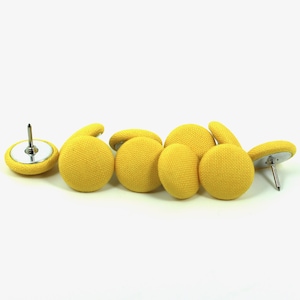 Op de afbeelding: Een verzameling gele stoffen bekledingsnagels. De nagels hebben een ronde, knoopachtige uitstraling met een zilverkleurige metalen basis en een scherpe pin. De stof heeft een getextureerde weefsel.