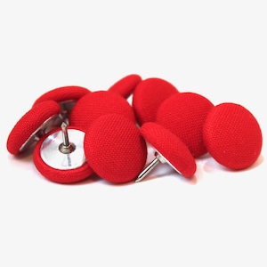 May include: A close-up of ten red fabric covered tacks with silver metal bases and points. The tacks are arranged in a pile on a white surface.