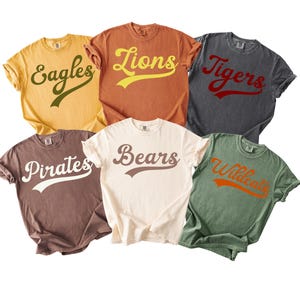 Puede incluir: Seis camisetas de manga corta en varios colores, cada una con el nombre de un equipo en una fuente retro. Las camisetas son en tonos de amarillo, naranja, carbón, marrón, crema y verde. Los nombres de los equipos incluyen Eagles, Lions, Tigers, Pirates, Bears y Wildcats.
