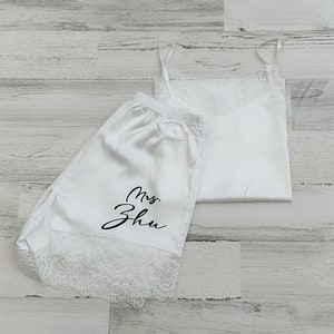 May include: White satin pajama shorts with lace trim and the text "Mrs. Zhu" in black script. The shorts are paired with a white satin tank top with lace trim.