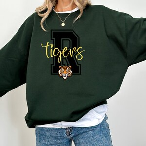 May include: A dark green sweatshirt with a black and white letter "R" and the word "tigers" in yellow lettering. A tiger face is inside the letter "R".