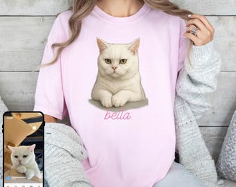 May include: A light pink short-sleeved t-shirt featuring a centered graphic of a white cat with yellow eyes and the name bella written in cursive pink script below the cat. The cat is shown in a seated position with its front paws tucked inward.