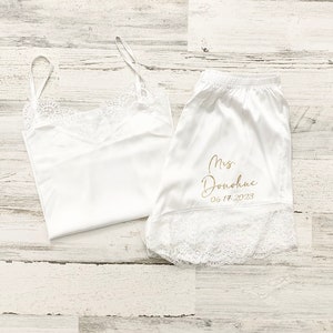 May include: White satin pajama set with lace trim. The top is a tank top style and the shorts have the text "Mrs. Donahue 06.17.2023" embroidered in gold.