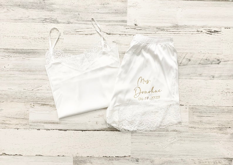 May include: White satin pajama set with lace trim. The top is a tank top style and the shorts have the text "Mrs. Donahue 06.17.2023" embroidered in gold.