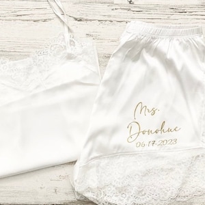 May include: White satin pajama set with lace trim. The top is a tank top and the bottoms are shorts. The shorts have the text "Mrs. Donohue 06.17.2023" printed in gold.