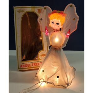 May include: A vintage illuminated angel tree topper with blonde hair, a pink bow, and white mesh wings. The figure wears a white mesh dress with sequins and holds two red electric candles. The original cardboard box is visible in the background.