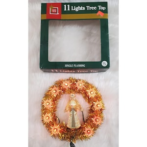May include: A Santa Trim 11 Lights Tree Top box with a rectangular cutout and a gold tinsel wreath featuring a central angel figure surrounded by eleven star-shaped light covers. The packaging text reads 11 Lights Tree Top and SINGLE FLASHING.