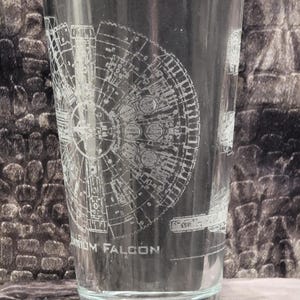 May include: A clear glass pint glass with an etched design of the Millennium Falcon spaceship. The text "Millennium Falcon" is etched at the bottom of the glass.