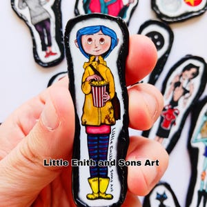 May include: A collection of handmade art pieces featuring characters from the animated film "Coraline." The central piece depicts a girl in a yellow raincoat holding popcorn. Other pieces show various characters. The text "Little Enith and Sons Art" is visible.