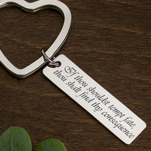 May include: A silver-colored heart-shaped keychain with a rectangular tag. The tag has the engraved quote, "If thou shouldst tempt fate, thou shalt find thy consequence." The keychain is on a dark wood surface.