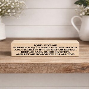 May include: A rectangular wooden block with black text. The text reads: "LORD, GIVE ME STRENGTH & COURAGE FOR THE MATCH, AND HUMILITY IN VICTORY OR DEFEAT. KEEP ME SAFE, GUIDE MY STEPS, AND LET ME HONOR YOU IN ALL I DO."