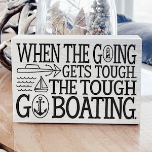 May include: A rectangular, white wooden sign with black text and nautical illustrations. The text reads "WHEN THE GOING GETS TOUGH, THE TOUGH GO BOATING." The sign features a boat, waves, a sailboat, an anchor, and a lighthouse.