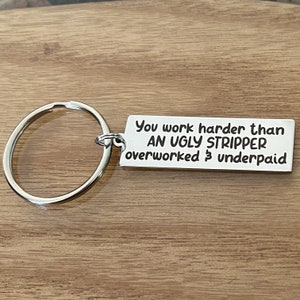 May include: A silver-colored keychain with a rectangular tag. The tag has the engraved text: "You work harder than AN UGLY STRIPPER overworked & underpaid". The keychain ring is large and circular.