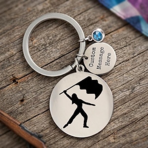 May include: Silver keychain with a round charm featuring a black silhouette of a person holding a flag. A smaller charm with a blue gem and a charm with the text "Custom Message Here" are also attached. The keychain is on a wooden surface.