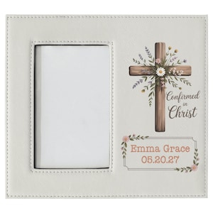 May include: Cream-colored photo frame with a rectangular opening and decorative cross design. The cross is adorned with flowers and the words "Confirmed in Christ." Below, it reads "Emma Grace 05.20.27."