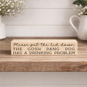May include: A rectangular wooden block with the text "Please put the lid down THE GOSH DANG DOG HAS A DRINKING PROBLEM" in black lettering. The block is on a wooden surface, with white vases and flowers in the background.