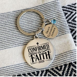 May include: A silver keychain with a large round pendant that reads "CONFIRMED IN FAITH 2026" in black text. A smaller pendant with the name "Grace Elizabeth Jansen" is attached, along with a blue gemstone. The keychain is on a striped fabric.