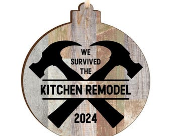 May include: A round wooden ornament with a weathered, multi-colored wood grain background. It features two black crossed hammers and the black text "WE SURVIVED THE KITCHEN REMODEL 2024" printed across the center of the design.