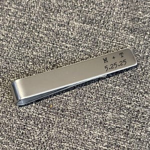 A silver metal binder clip with a personalized message engraved on it, resting on a textured gray surface.