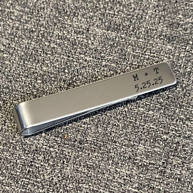 A silver metal binder clip with a personalized message engraved on it, resting on a textured gray surface.
