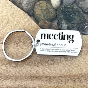 May include: A white rectangular keychain with the word "meeting" in bold black font. The keychain includes a definition of "meeting" and a silver metal key ring. The background is a wooden surface.