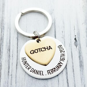 May include: A silver-toned circular keychain featuring a smaller gold-toned heart-shaped charm attached to the ring. The heart is engraved with the word GOTCHA, while the circular disc is engraved with HUNTER DANIEL and FEBRUARY 15TH 2019.