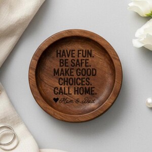 May include: A round, wooden trinket dish with a dark brown finish. The dish features engraved text that reads "HAVE FUN. BE SAFE. MAKE GOOD CHOICES. CALL HOME." with "Mom & Dad" below. A heart symbol is included.