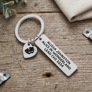May include: A silver keychain with a heart-shaped charm featuring theater masks and a rectangular tag. The tag has the text "ALISSA JOHNSTON, ALICE IN WONDERLAND, LEAD CHS 2024" in black. The keychain is on a wooden surface.