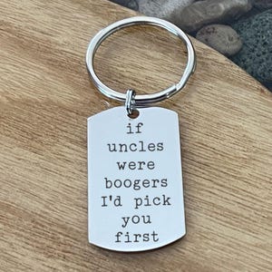 May include: A silver-colored metal keychain with a rectangular tag. The tag has the text "if uncles were boogers I'd pick you first" in a simple font. The keychain is on a wooden surface.