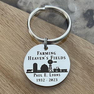 May include: A silver-colored keychain with a round pendant featuring the text "Farming Heaven's Fields" and a farm scene silhouette. The keychain includes a cow, silo, barn, windmill, tractor, and the name "Paul E. Lyons 1932-2023".