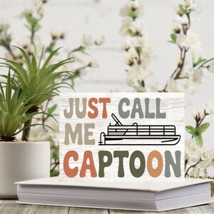 May include: A wooden sign with the text "JUST CALL ME CAPTOON" in various colors, with a black and white illustration of a pontoon boat. The sign is placed on a white book, with a small potted plant and floral arrangement in the background.