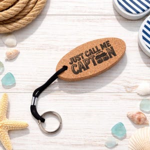 May include: An oval cork keychain with the text "JUST CALL ME CAPT'N" and a boat graphic. The keychain has a black cord and a silver key ring. The background includes seashells, sea glass, and rope.